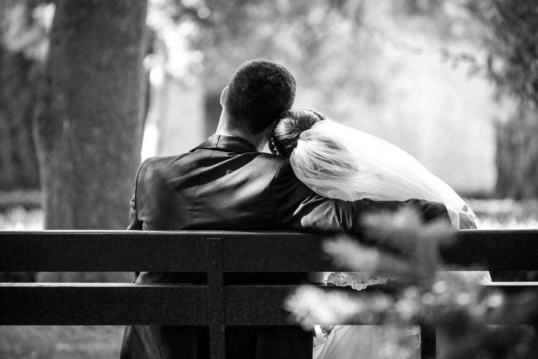 Fotografie alb-negru cu mireasa care isi sprijina capul pe umarul mirelui, pe banca, surprinsi din spate de fotograful de nunta la sedinta foto in parc