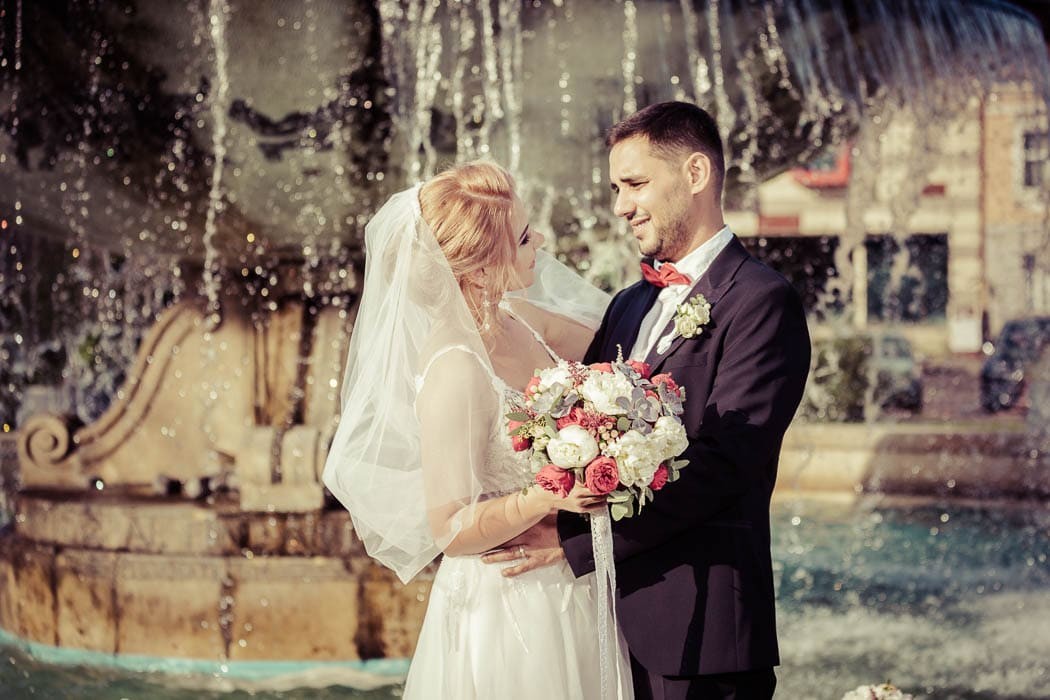 Mirele si mireasa se privesc cu dragoste, in fata unei fantai decorative cu apa, la sedinta foto de nunta
