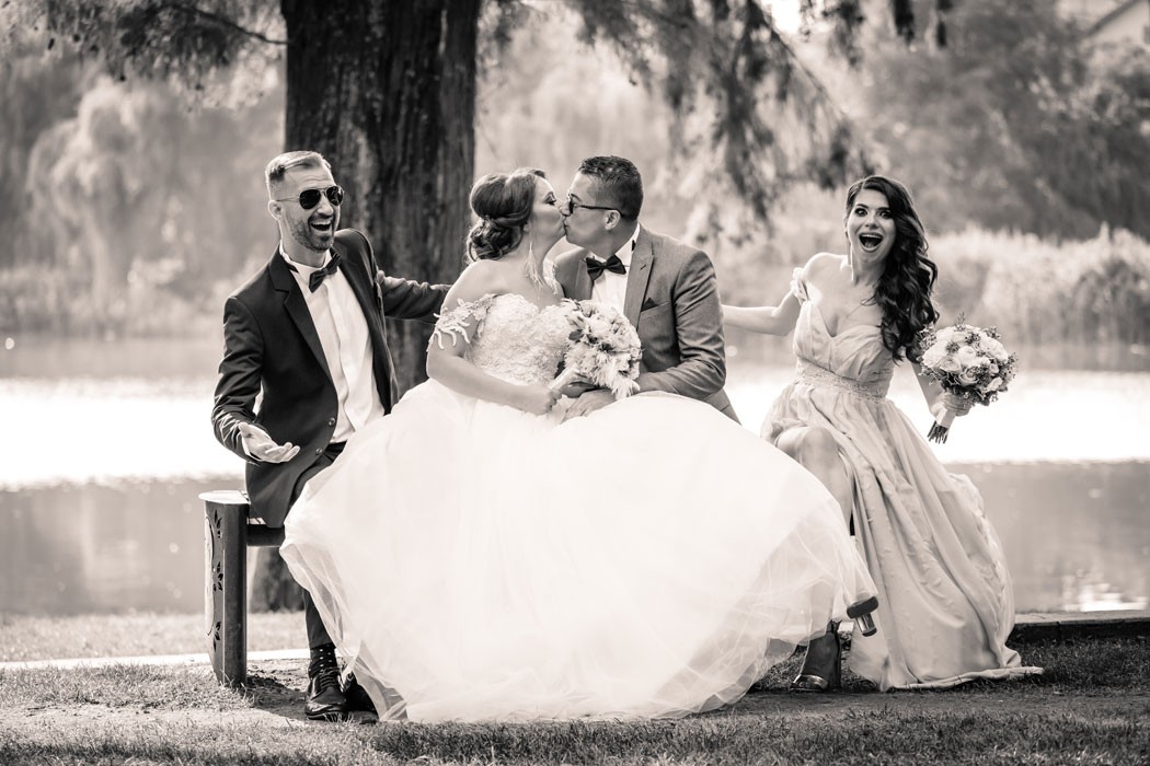 Fotografie editata in sepia, in parc pe banca stau mirii si nasii, mirele saruta mireasa, in stanga este nasul care se bucura iar in dreapta nasa se mira