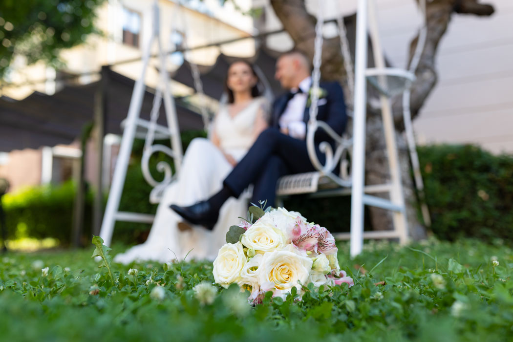 Fotografie de nunta cu efectul bokeh aplicat, buchetul miresei pe iarba in prim plan in clar si mirii in fundalul blurat pe un balansoar