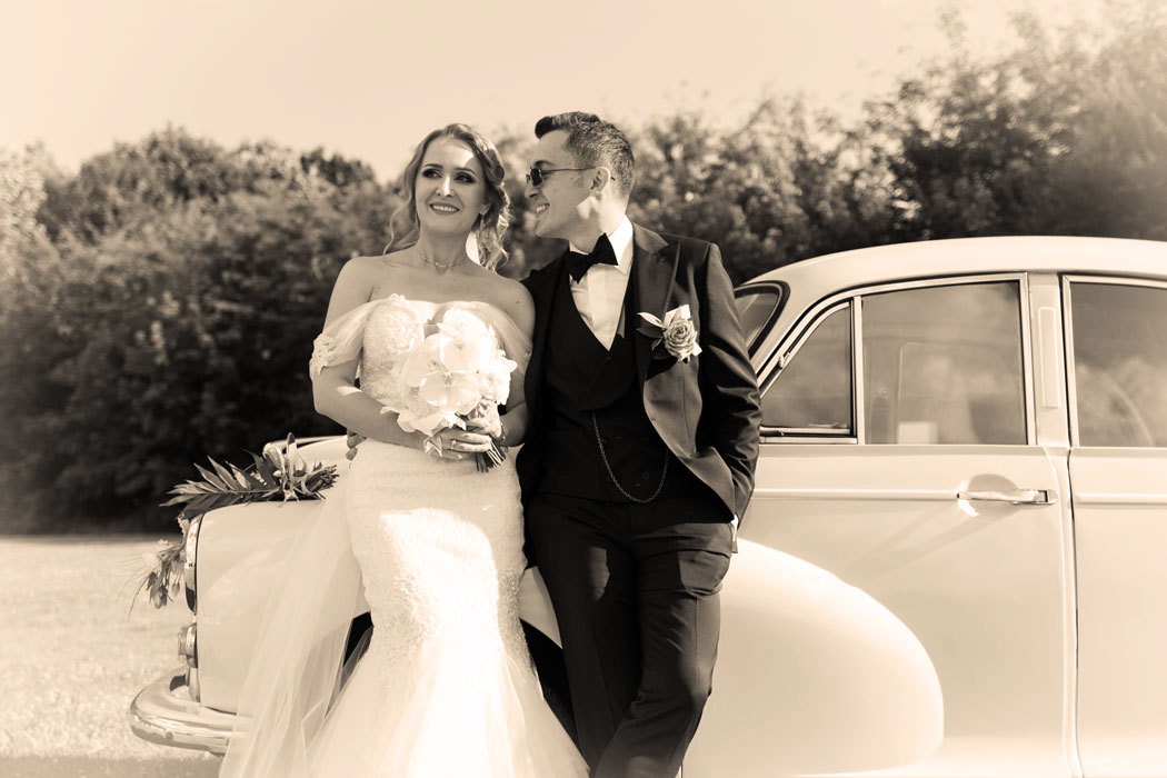 Fotografie de nunta cu efect sepia aplicat, mirele si mireasa la sedinta foto stau unul langa altul sprijiniti de un automobil vintage