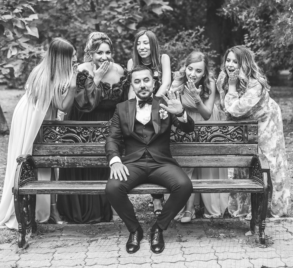 Fotografie alb-negru cu mirele care se uita la verigheta de pe mana pe banca in parc, insotit de domnisoarele de onoare si nasa, cadru surprins de fotograf la sedinta foto de nunta in Bucuresti