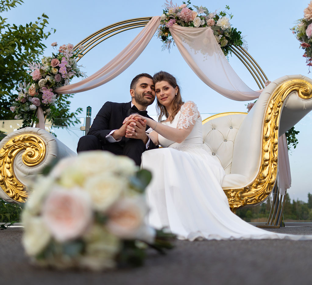 Mire si mireasa tinandu-se de mana pe o canapea alba cu accente aurii sub o arcada florala, la sedinta foto de nunta in Bucuresti