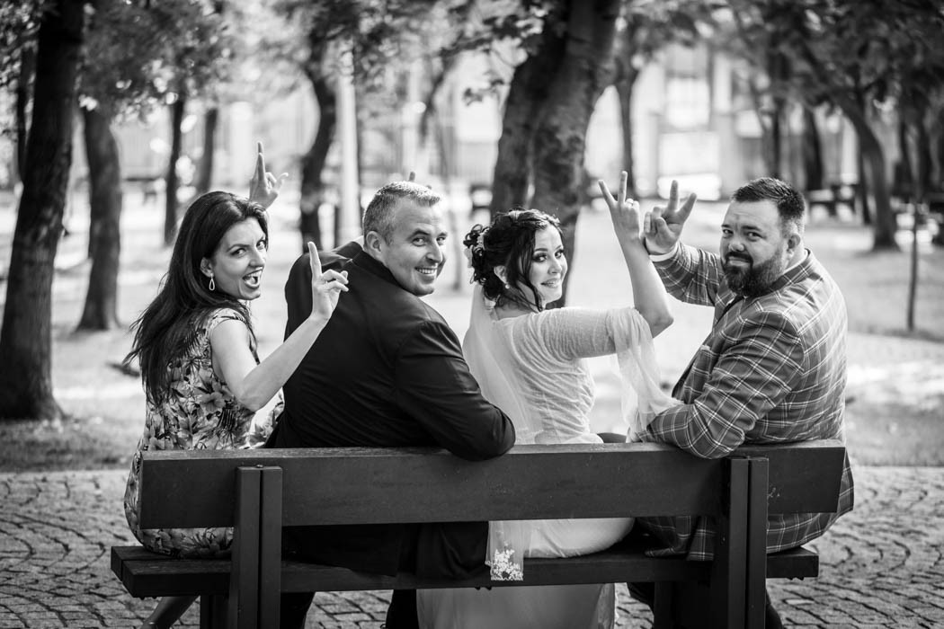 Fotografie monocroma alb-negru cu mirii si nasii pe o banca in parc, afisand semnul victoriei, intorsi cu fata la fotograful de nunta