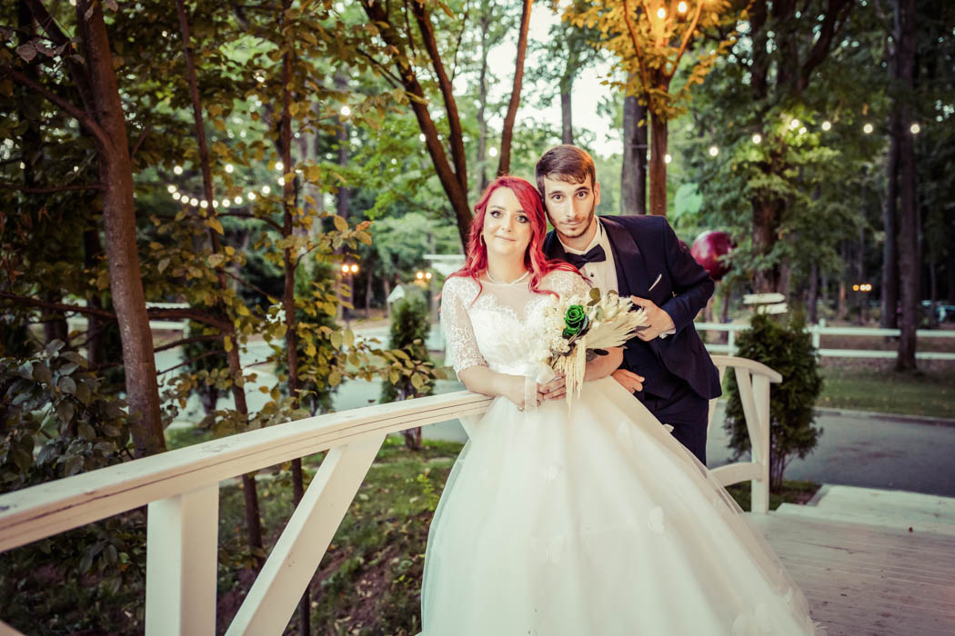 Mirele langa mireasa pe un pod de lemn, la sedinta foto de nunta in gradina cu ghirlande de lumini, buchetul de flori in mainile miresei