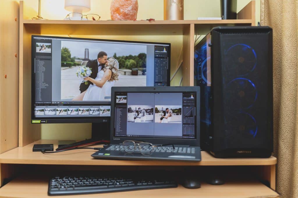 Birou de calculator cu sertar pe care se afla echipamentele de editare foto: PC, laptop, monitor cu diagonala mare, tastatura si mouse, pe ecrane se vad fotografiile in etapa de procesare din Lightroom