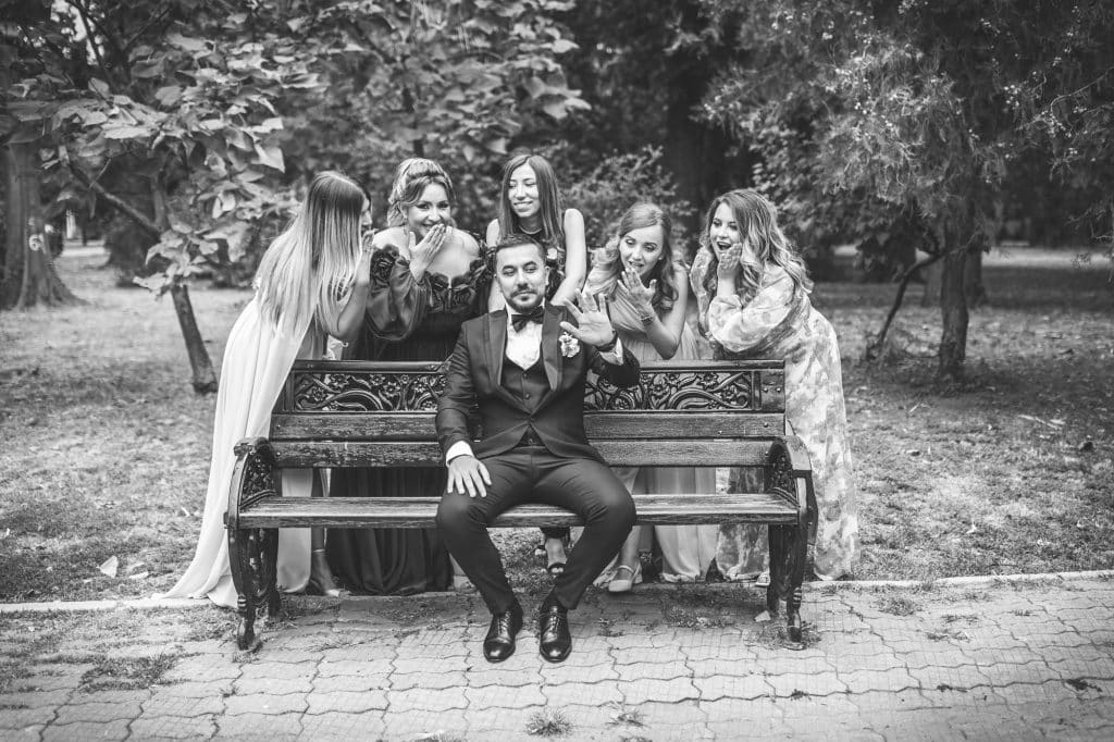 Fotografie alb-negru la sedinta foto de nunta, mirele pe banca in parc se uita la verigheta de pe mana stanga ridicata, nasa si domnisoarele de onoare in picioare in spatele bancii se mira