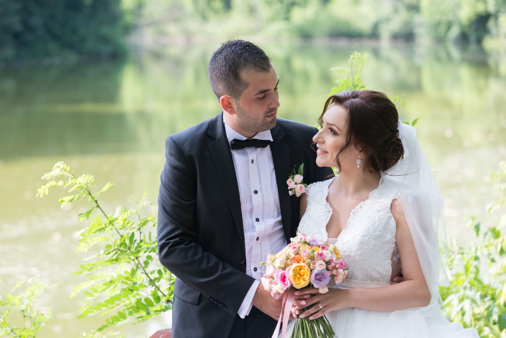Mire si mireasa la sedinta foto de nunta, privind unul spre celalalt langa un lac, mireasa tinand buchetul de flori