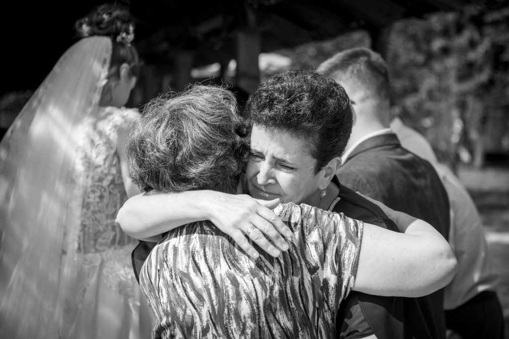 Fotografie spontana cu mama mirelui si mama miresei imbratisandu-se cu lacrimi in ochi, in fundal blur sunt mirele si mireasa inaintea ceremoniei religioase